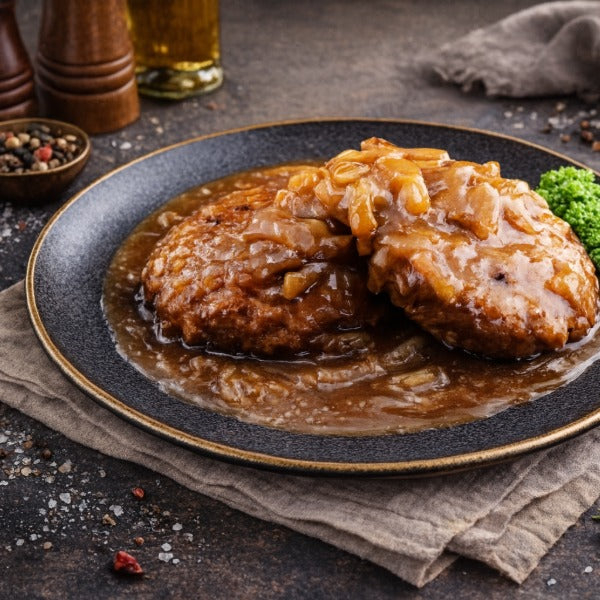 Zwei gebratene Hacksteaks in brauner Zwiebelsauce auf dunklem Teller, garniert mit etwas Brokkoli, auf rustikalem Untergrund mit Gewürzen im Hintergrund.“