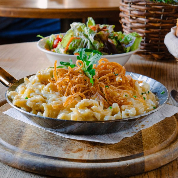 Pfanne mit Käsespätzle und knusprigen Röstzwiebeln, garniert mit Petersilie, im Hintergrund ein gemischter Salat auf rustikalem Holztisch.