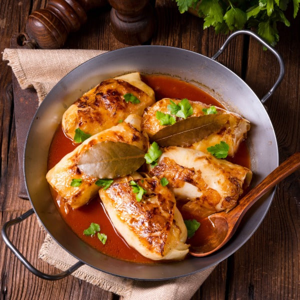 Goldbraun geschmorte Kohlrouladen in Tomatensauce mit Lorbeerblättern und Petersilie in einer Pfanne, dazu ein Holzlöffel auf rustikalem Holztisch.
