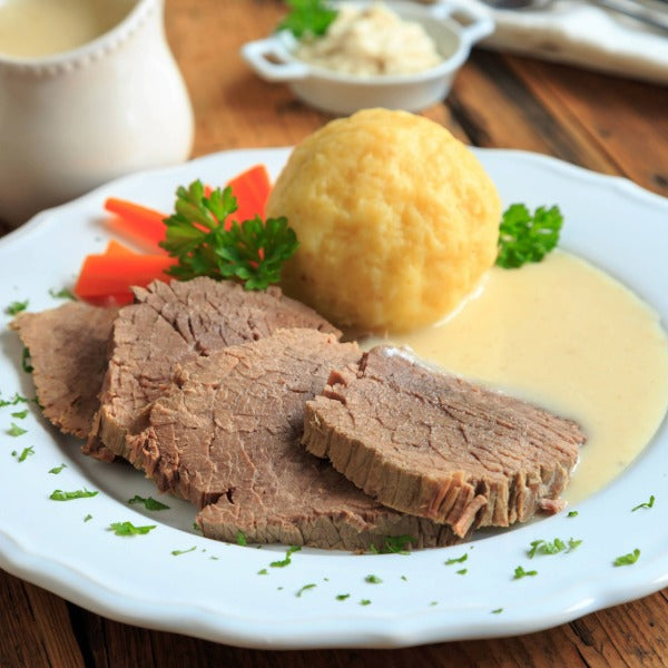 Gekochtes Rindfleisch in heller Meerrettichsauce mit Kartoffelkl0ß und Möhrenstiften, angerichtet auf weißem Teller auf rustikalem Holztisch.