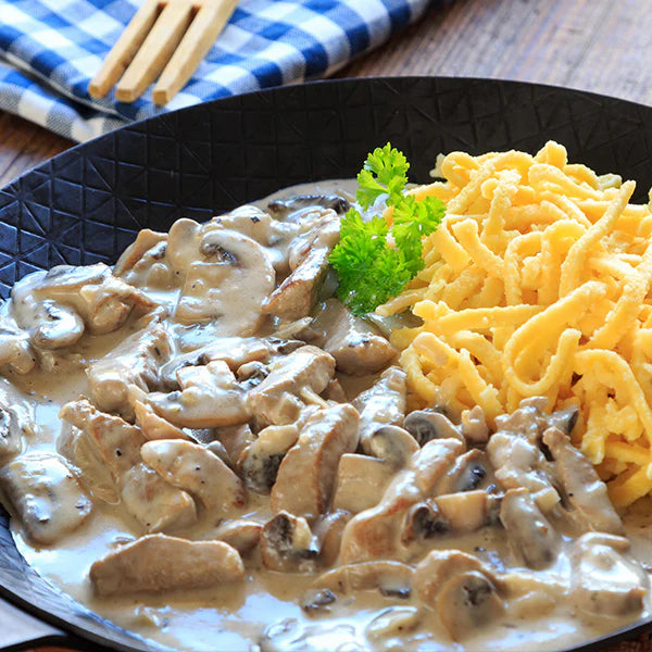 Schweinegeschnetzeltes in Champignon-Rahmsauce, serviert mit Spätzle und garniert mit frischer Petersilie in einer schwarzen Pfanne