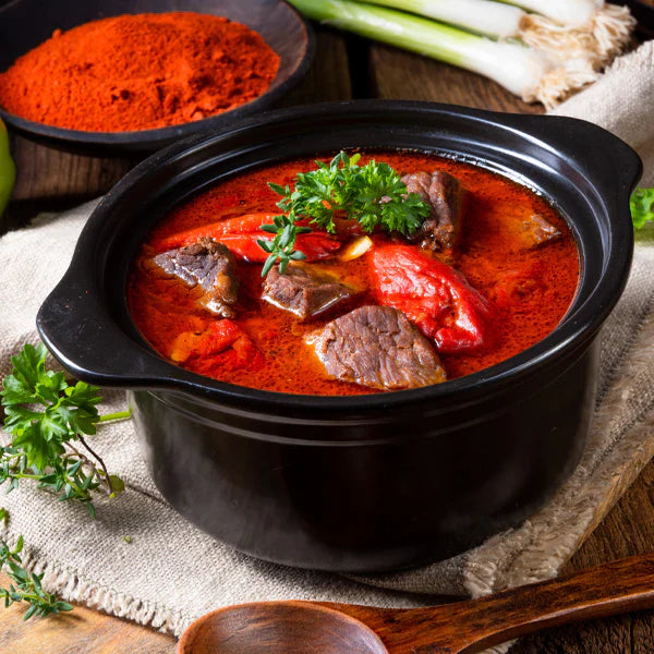 Rustikaler Schaschliktopf mit Stücken von Rindfleisch und Paprika in roter Paprikasauce, serviert im schwarzen Topf und garniert mit frischer Petersilie auf Holztisch.