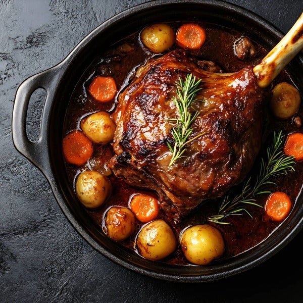 Geschmorte Haxe mit knuspriger Kruste in dunkler Bratensauce, umgeben von kleinen Kartoffeln und Möhren, garniert mit Rosmarin im gusseisernen Bräter auf dunklem Hintergrund.
