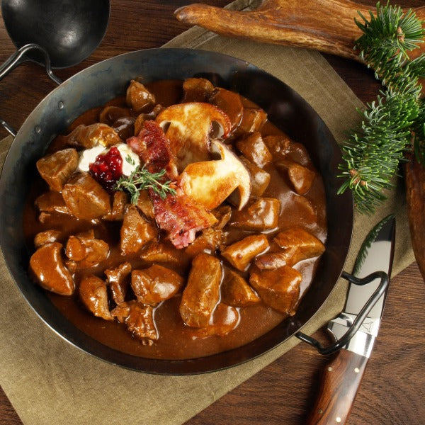 Herzhaftes Hirschgulasch in dunkler Sauce in einer Pfanne, garniert mit Pilz, Speck, Preiselbeeren und Kräutern, dekoriert mit Tannenzweig und Jagdmesser auf rustikalem Holztisch.