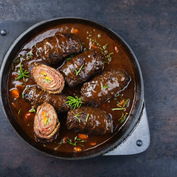 Rinderrouladen in dunkler Bratensauce in einer Pfanne, teilweise aufgeschnitten mit sichtbarer Füllung, garniert mit frischem Rosmarin auf dunklem Untergrund.