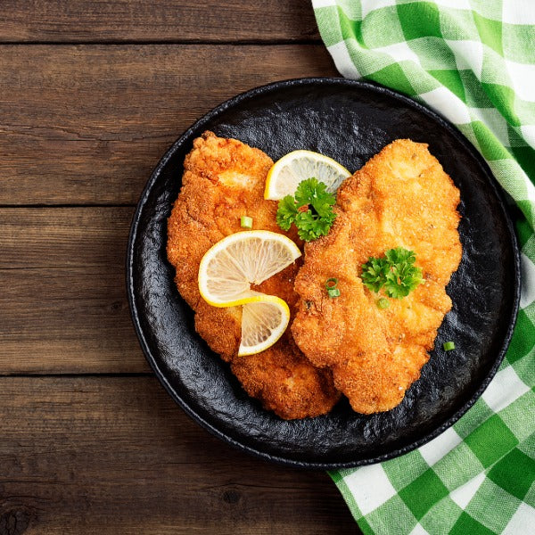 „Zwei goldbraun panierte Schnitzel mit Zitronenscheiben und Petersilie auf schwarzem Teller, daneben ein grün-weiß kariertes Tuch auf rustikalem Holztisch.