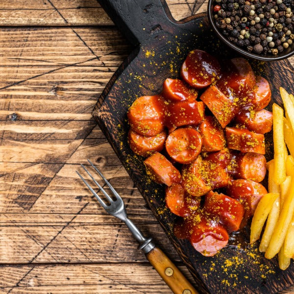 Geschnittene Currywurst mit roter Currysauce und Pommes auf einem dunklen Holzbrett, daneben eine kleine Gabel und ein Schälchen Pfefferkörner auf rustikalem Holztisch.