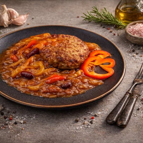 Gebratenes Hacksteak auf Paprika-Zwiebelsauce mit Bohnen, garniert mit roten Paprikaringen auf dunklem Teller, daneben Besteck, Rosmarinzweig und Gewürze auf steinigem Untergrund.