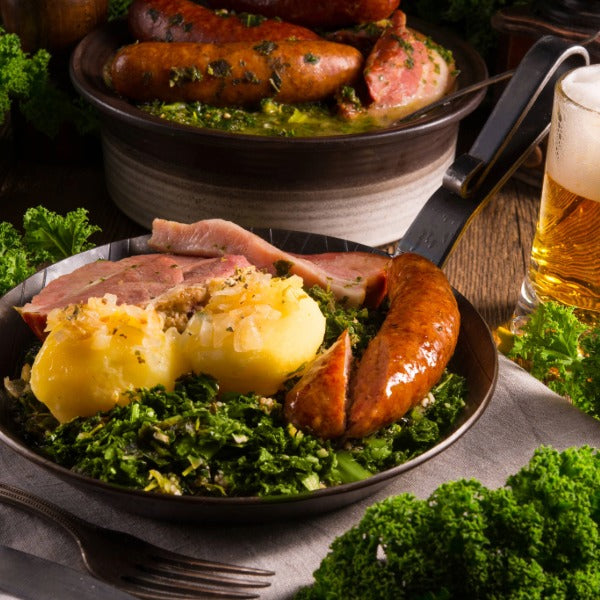 Deftiges Grünkohlgericht mit Kartoffeln, geräuchertem Kasseler und Wurst auf einem Teller, im Hintergrund weitere Wurstpfanne und Glas Bier auf rustikalem Holztisch.