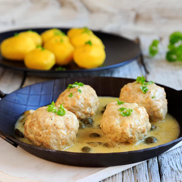 Königsberger Klopse in einer schwarzen Pfanne, serviert mit Kapernsauce und Kartoffeln im Hintergrund